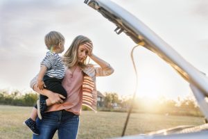 Portrait of young Caucasian woman standing by broken down car and waiting for assistance while holding her baby boy during sunset. Photo of female waits for assistance near her car broken down on the road side. Frustrated young brown hair woman looking at broken down car engine on street. Woman does not understand why her car engine failed. Desperate mother holding her angry baby.