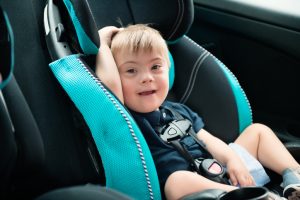 Little boy with Down Syndrome is buckled into car seat. He is smiling and looking the camera. He has blond hair et blue eyes. He wear blue short and sweater. Color and horizontal. Smiling boy with Down Syndrome is buckled into car seat.
