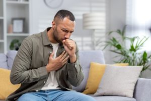 Young African American man sitting on the couch at home and coughing. Suffers from an attack of asthma, allergies. He holds his chest, covers his mouth with his hand. inhalation, smoke inhalation, inhalation injury, work injury, child injury, personal injury.